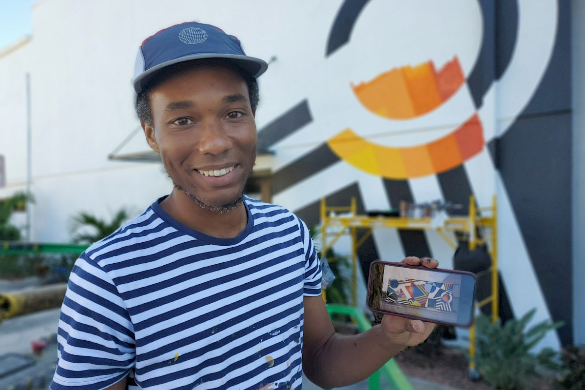 A person in a striped shirt and hat is smiling while holding a phone; there's a colorful mural in the background.
