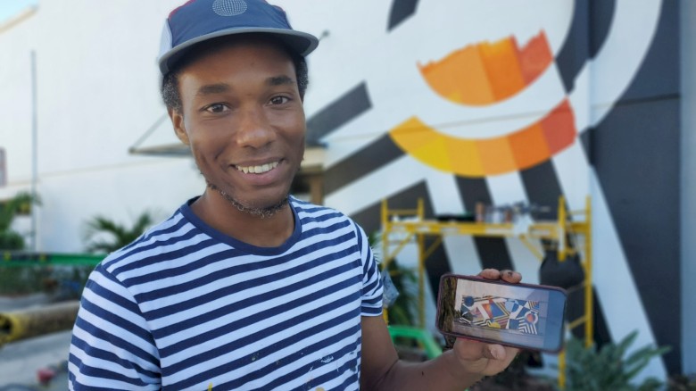 A person in a striped shirt and hat is smiling while holding a phone; there's a colorful mural in the background.