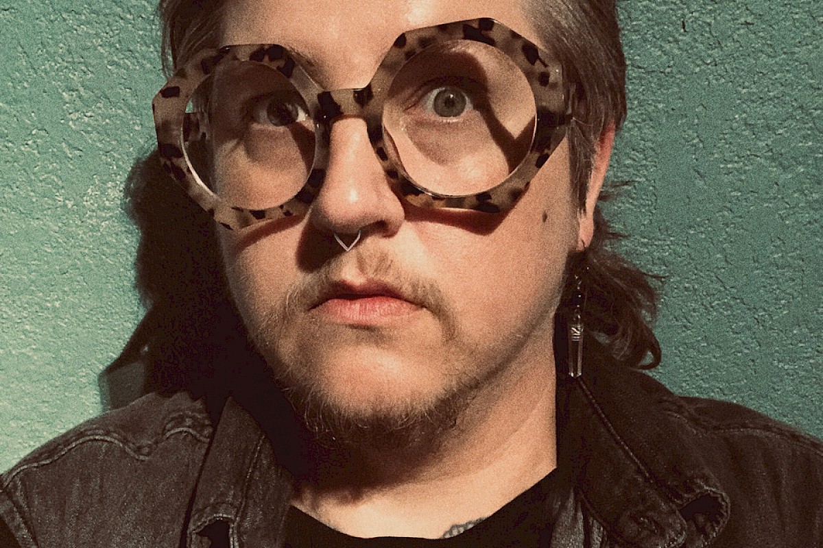 A person is wearing large, round, tortoiseshell glasses, a black shirt, and a denim jacket. They have short hair and are posing against a green wall.