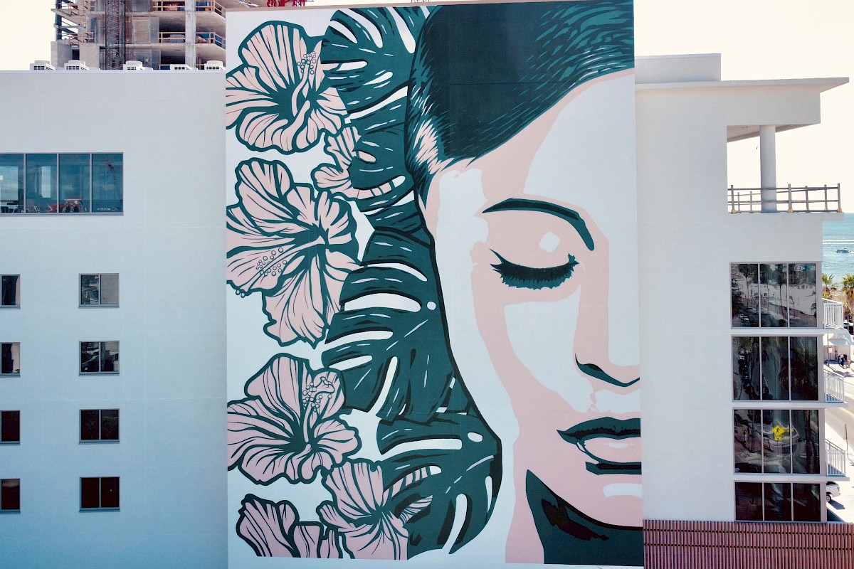 A large mural of a serene woman's face with closed eyes and floral designs is painted on the side of a modern white building near the waterfront.