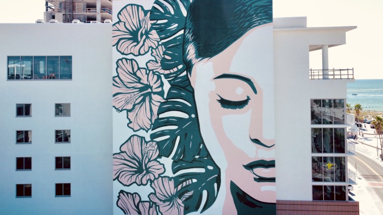 A large mural of a serene woman's face with closed eyes and floral designs is painted on the side of a modern white building near the waterfront.