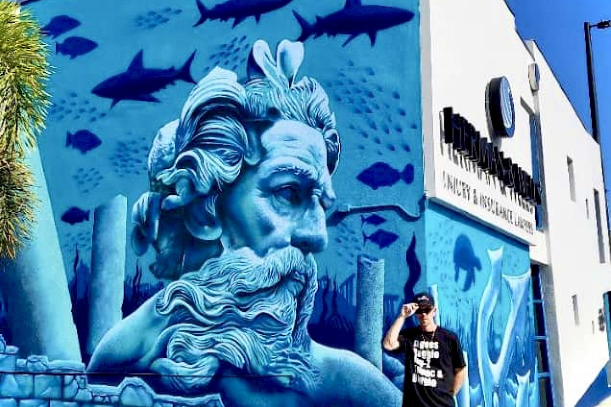 A person stands next to a vibrant mural depicting an underwater scene with a large, blue-toned face, fish, and sharks on a building.