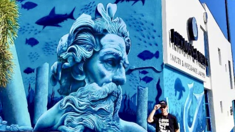 A person stands next to a vibrant mural depicting an underwater scene with a large, blue-toned face, fish, and sharks on a building.