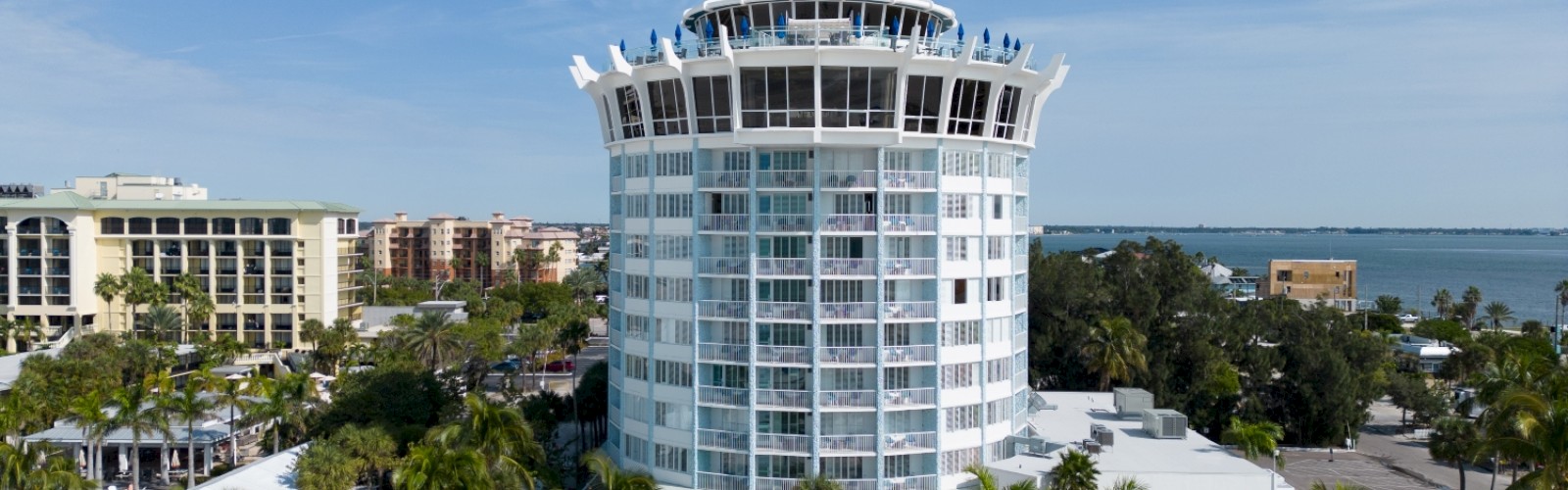 A tall, cylindrical building by the beach, surrounded by palm trees and neighboring structures, under a partly cloudy blue sky, ending the sentence.