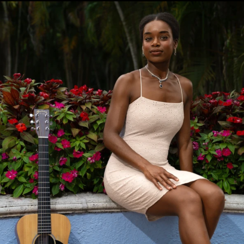 Kim sits by colorful flowers and greenery next to a guitar, exuding elegance in an outdoor setting.