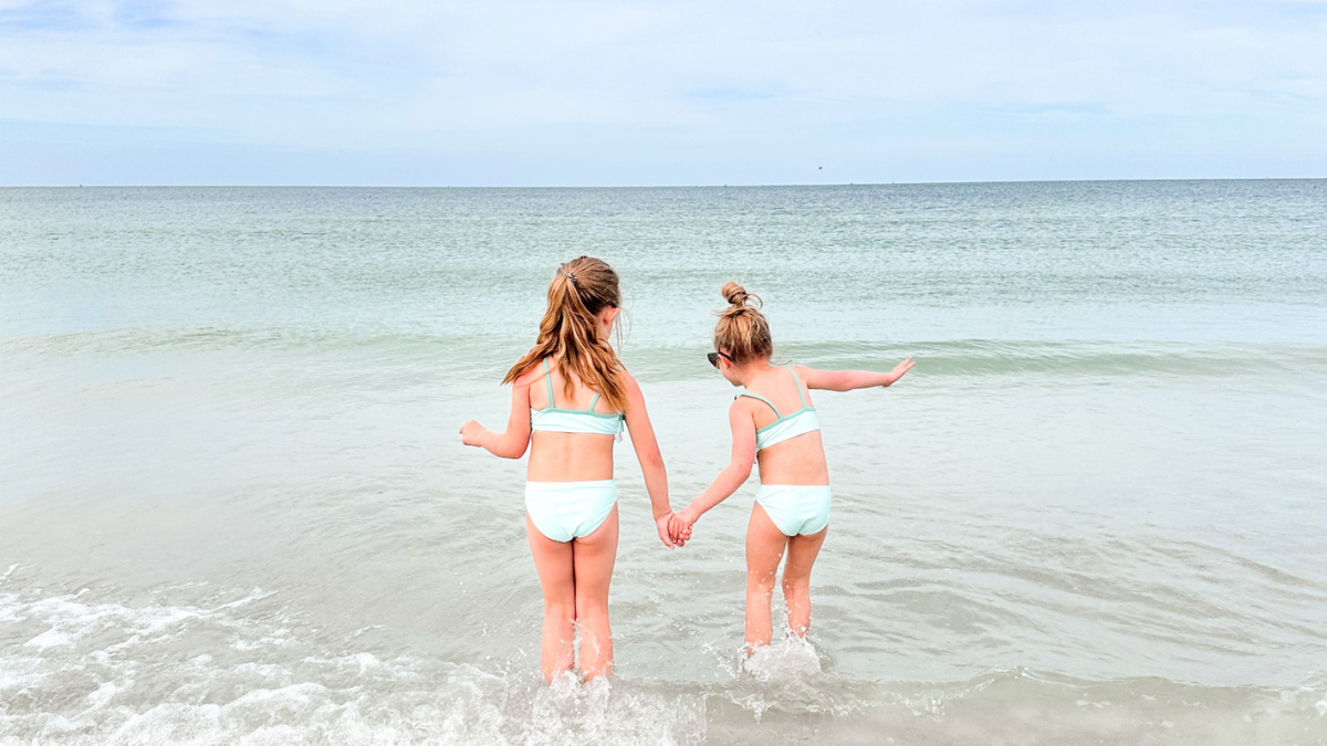 Two children in swimsuits holding hands and playing at the edge of the ocean on a sandy beach under a cloudy sky.