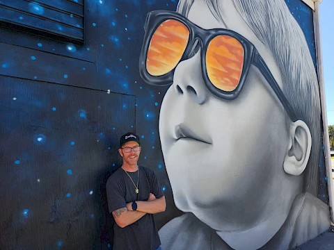 A man stands next to a wall mural of a child wearing sunglasses reflecting an orange sky, with a dark, starry background in the mural.