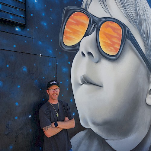 A man stands next to a wall mural of a child wearing sunglasses reflecting an orange sky, with a dark, starry background in the mural.