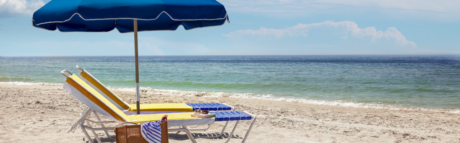 A sunny beach scene with two lounge chairs and a blue umbrella near the shoreline. A bag and striped towel are also visible on the chairs.