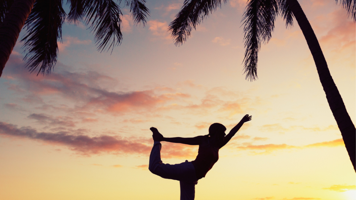 A person doing a yoga pose on a beach at sunset, flanked by palm trees and a vibrant sky.