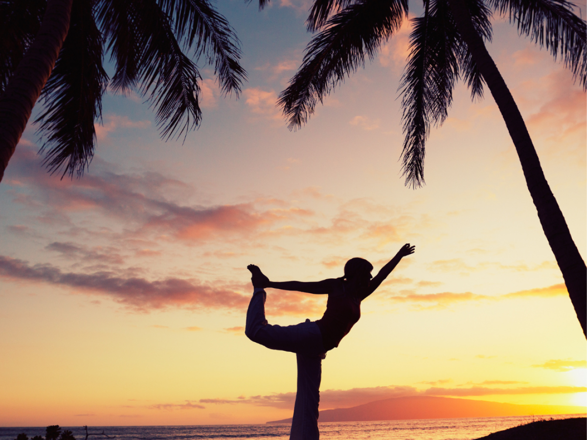 A person doing a yoga pose on a beach at sunset, flanked by palm trees and a vibrant sky.