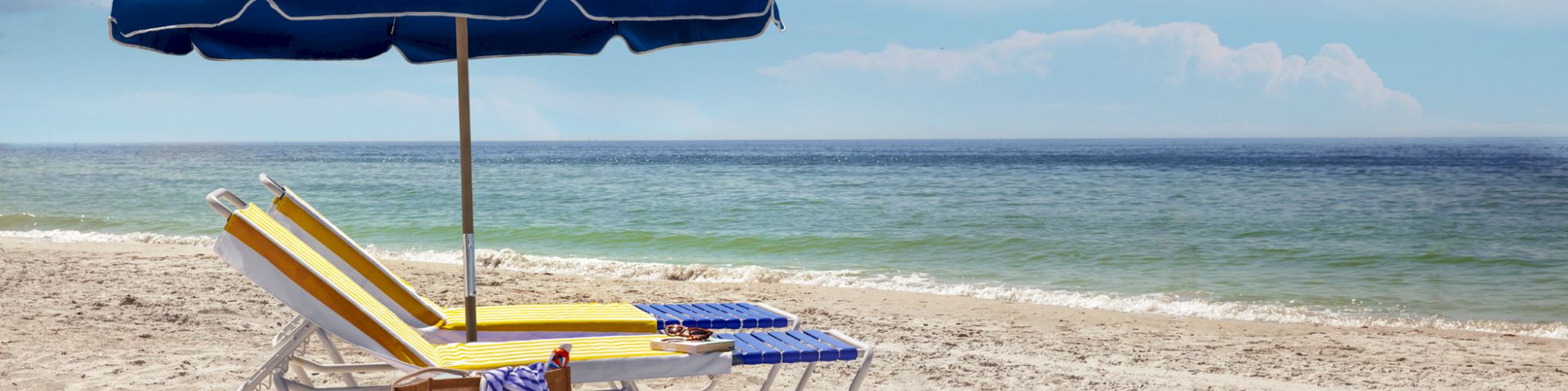 A sunny beach scene with two lounge chairs and a blue umbrella near the shoreline. A bag and striped towel are also visible on the chairs.