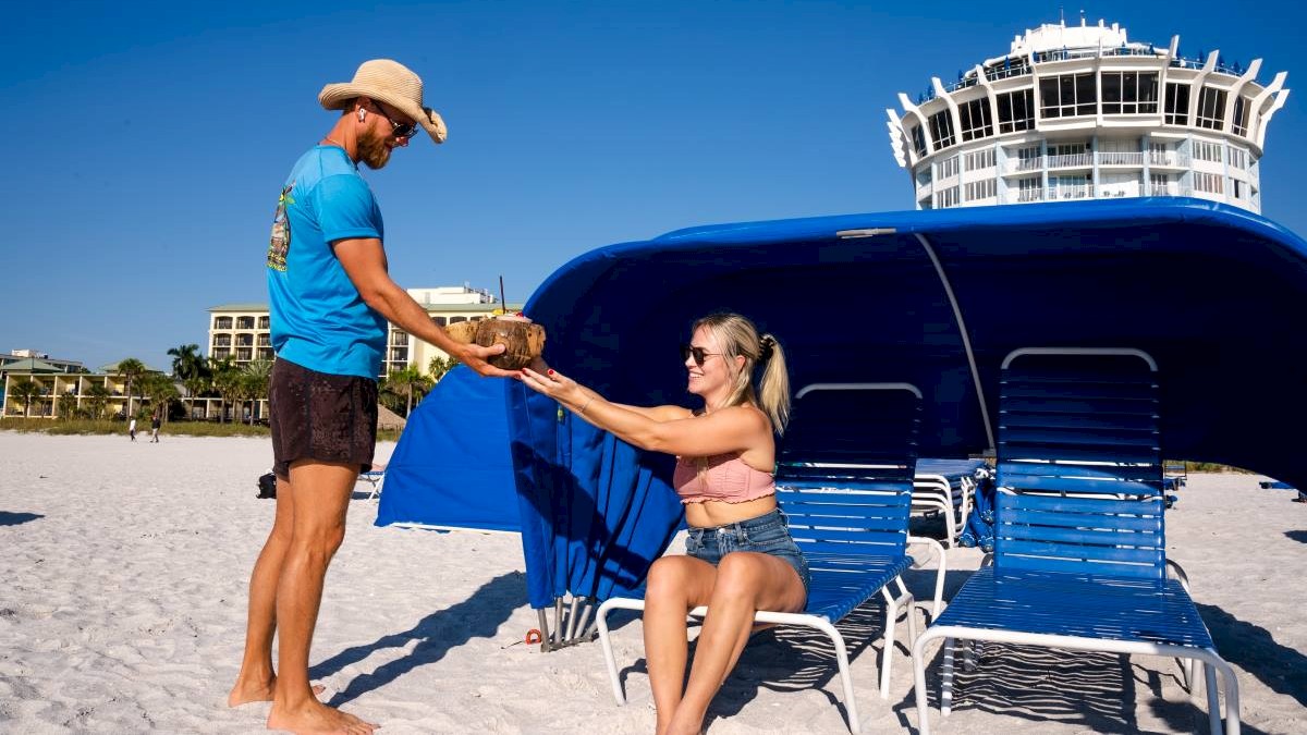 A man in a hat hands a coconut to a woman seated on a lounge chair under a blue canopy at the beach, with a building in the background.