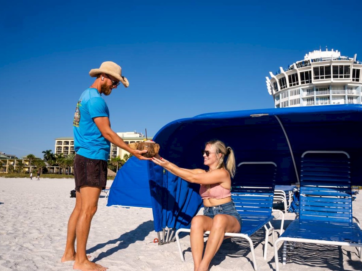 A man in a hat hands a coconut to a woman seated on a lounge chair under a blue canopy at the beach, with a building in the background.