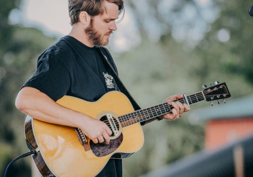 A person is playing an acoustic guitar outdoors on a stage with trees and blurred background elements visible.