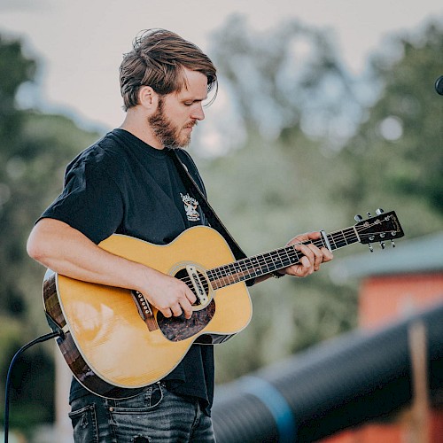 A person is playing an acoustic guitar outdoors on a stage with trees and blurred background elements visible.