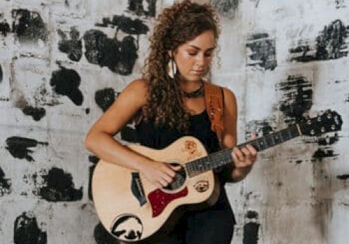 A woman with curly hair stands in front of a black and white painted wall, playing an acoustic guitar while wearing a black outfit.