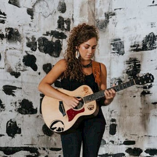 A woman with curly hair stands in front of a black and white painted wall, playing an acoustic guitar while wearing a black outfit.