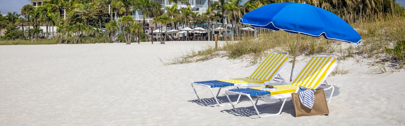 A sandy beach with two lounge chairs and a blue umbrella, in front of a modern, round high-rise building, surrounded by palm trees and clear skies.
