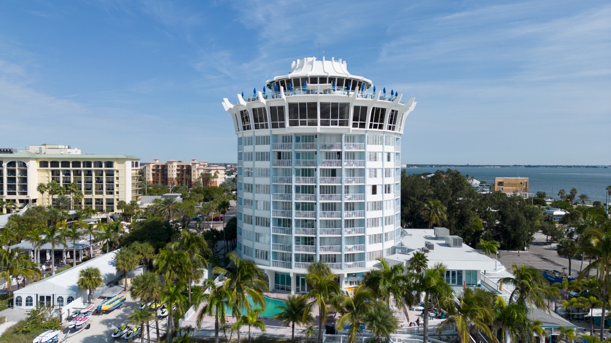 A modern, round, multi-story hotel with a unique rooftop design, surrounded by palm trees, near a body of water, on a clear, sunny day.