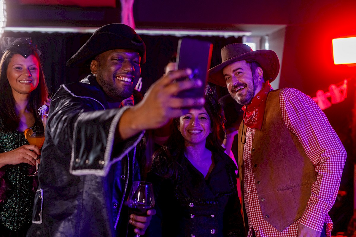 People in costumes taking a group selfie at a party. Two women and two men are smiling with drinks in hand, enjoying the celebration.