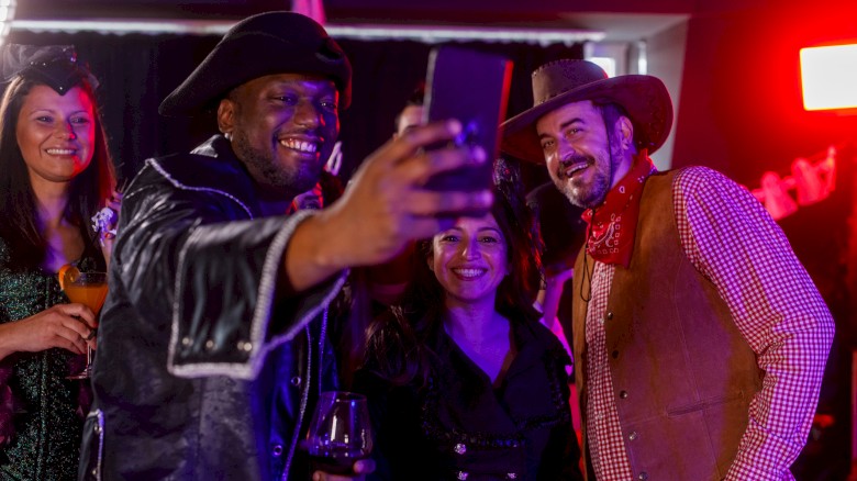 People in costumes taking a group selfie at a party. Two women and two men are smiling with drinks in hand, enjoying the celebration.