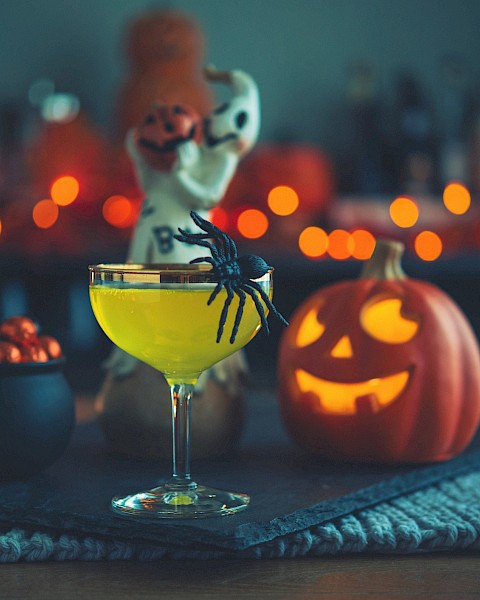 A Halloween-themed setup with a cocktail, a fake spider, a carved pumpkin, a small cauldron with chocolates, and spooky decorations in the background.