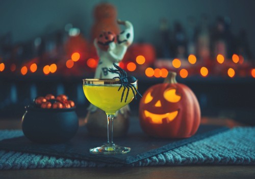 A Halloween-themed setup with a cocktail, a fake spider, a carved pumpkin, a small cauldron with chocolates, and spooky decorations in the background.