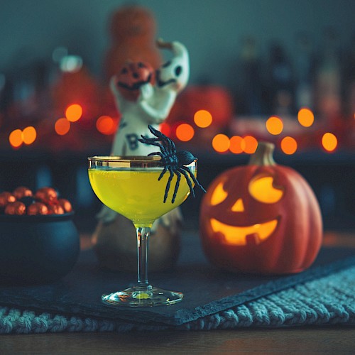 A Halloween-themed setup with a cocktail, a fake spider, a carved pumpkin, a small cauldron with chocolates, and spooky decorations in the background.