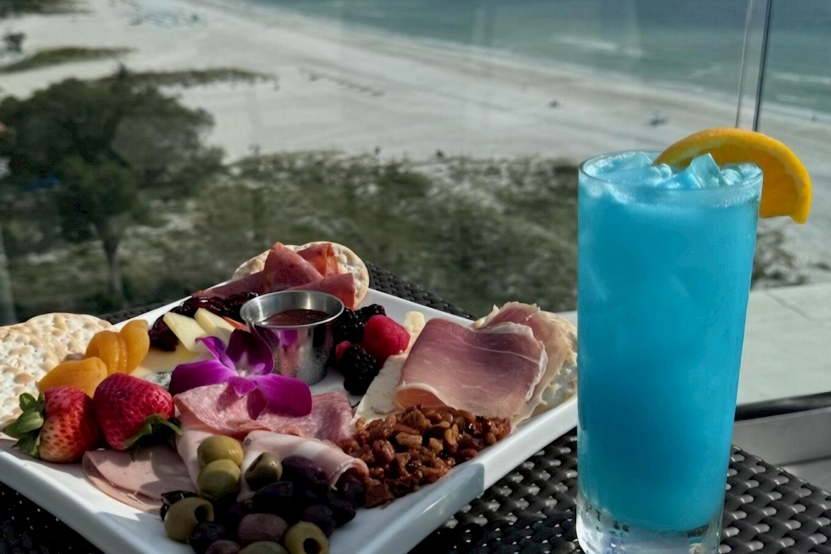 A charcuterie board and a blue cocktail with a lemon slice, set on an outdoor table overlooking a beautiful beach and ocean.