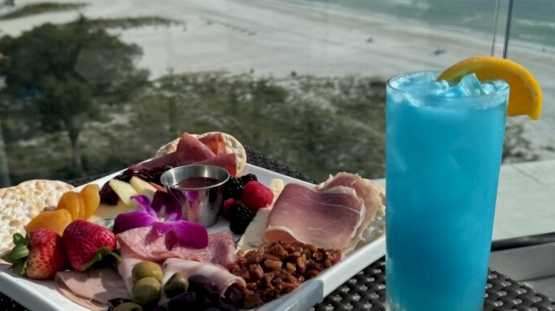 A charcuterie board and a blue cocktail with a lemon slice, set on an outdoor table overlooking a beautiful beach and ocean.