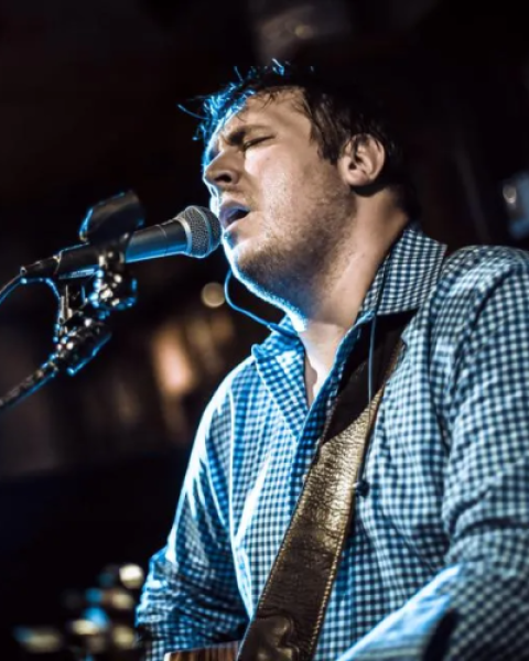 A person is passionately singing and playing a guitar into a microphone on a stand, wearing a checkered shirt in a dimly lit setting.