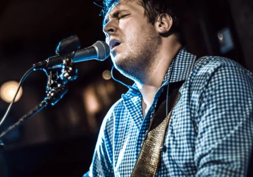 A person is passionately singing and playing a guitar into a microphone on a stand, wearing a checkered shirt in a dimly lit setting.