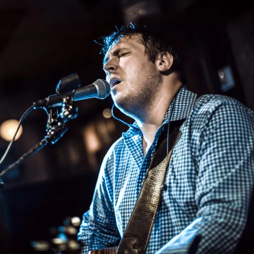 A person is passionately singing and playing a guitar into a microphone on a stand, wearing a checkered shirt in a dimly lit setting.