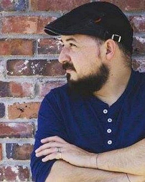 A man with a beard, wearing a blue shirt and a cap, is standing with his arms crossed, looking to his left, against a brick wall.