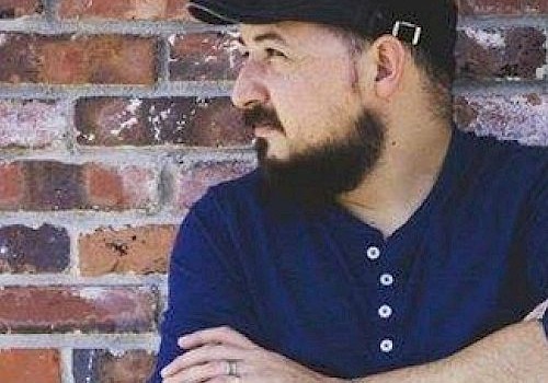 A man with a beard, wearing a blue shirt and a cap, is standing with his arms crossed, looking to his left, against a brick wall.