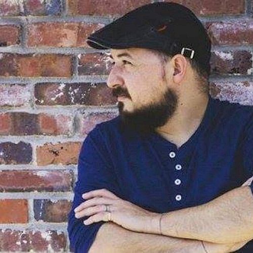 A man with a beard, wearing a blue shirt and a cap, is standing with his arms crossed, looking to his left, against a brick wall.