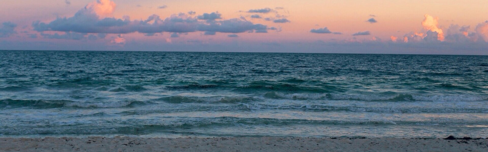 An image of a beach during sunset with the ocean waves gently meeting the sandy shore and a sky filled with soft pink and purple clouds.