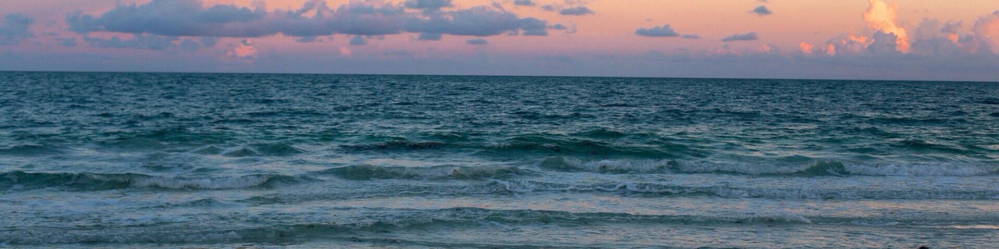 An image of a beach during sunset with the ocean waves gently meeting the sandy shore and a sky filled with soft pink and purple clouds.