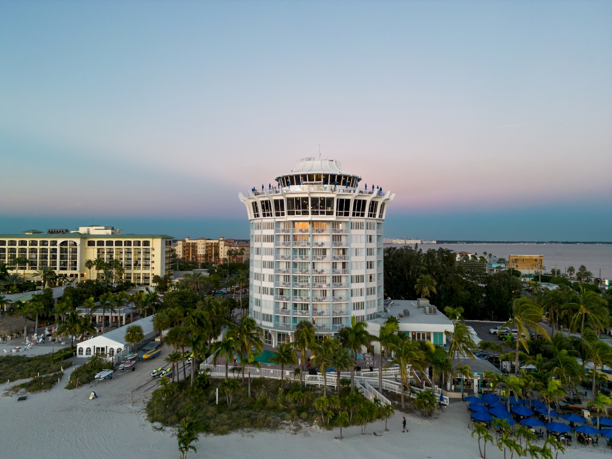 A round, multi-story building is situated on a beach with palm trees, surrounded by other structures, under a serene sky at dusk.