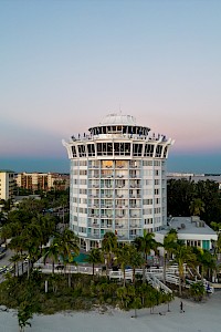 The image shows a tall, round building on a beach surrounded by palm trees, with additional buildings and the ocean in the background.